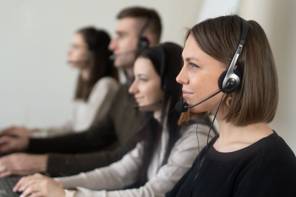 Side view of young coworkers in casual clothes sitting in light workplace with headphones with microphone while surfing on laptop